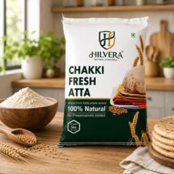 Hilvera Chakki Fresh Atta 5kg pack displayed on a wooden kitchen table next to a bowl of whole wheat flour and fresh rotis.