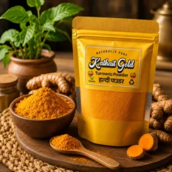 A golden stand-up pouch of Kathait Gold Turmeric Powder (Haldi Powder) with a clear window showing the rich yellow spice. The pouch sits on a rustic wooden board surrounded by a bowl of ground turmeric, a loaded wooden spoon, sliced turmeric roots, and a green potted plant.